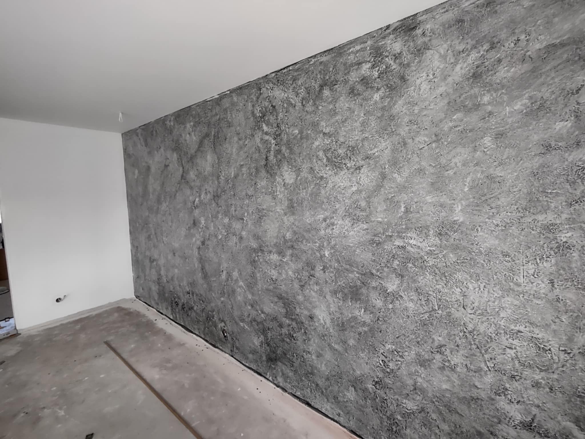 Ściana z efektem dekoracyjnym w odcieniach szarości, surowa faktura, widoczne nierówności i cieniowania. Podłoga surowa, przygotowana do dalszych prac wykończeniowych.