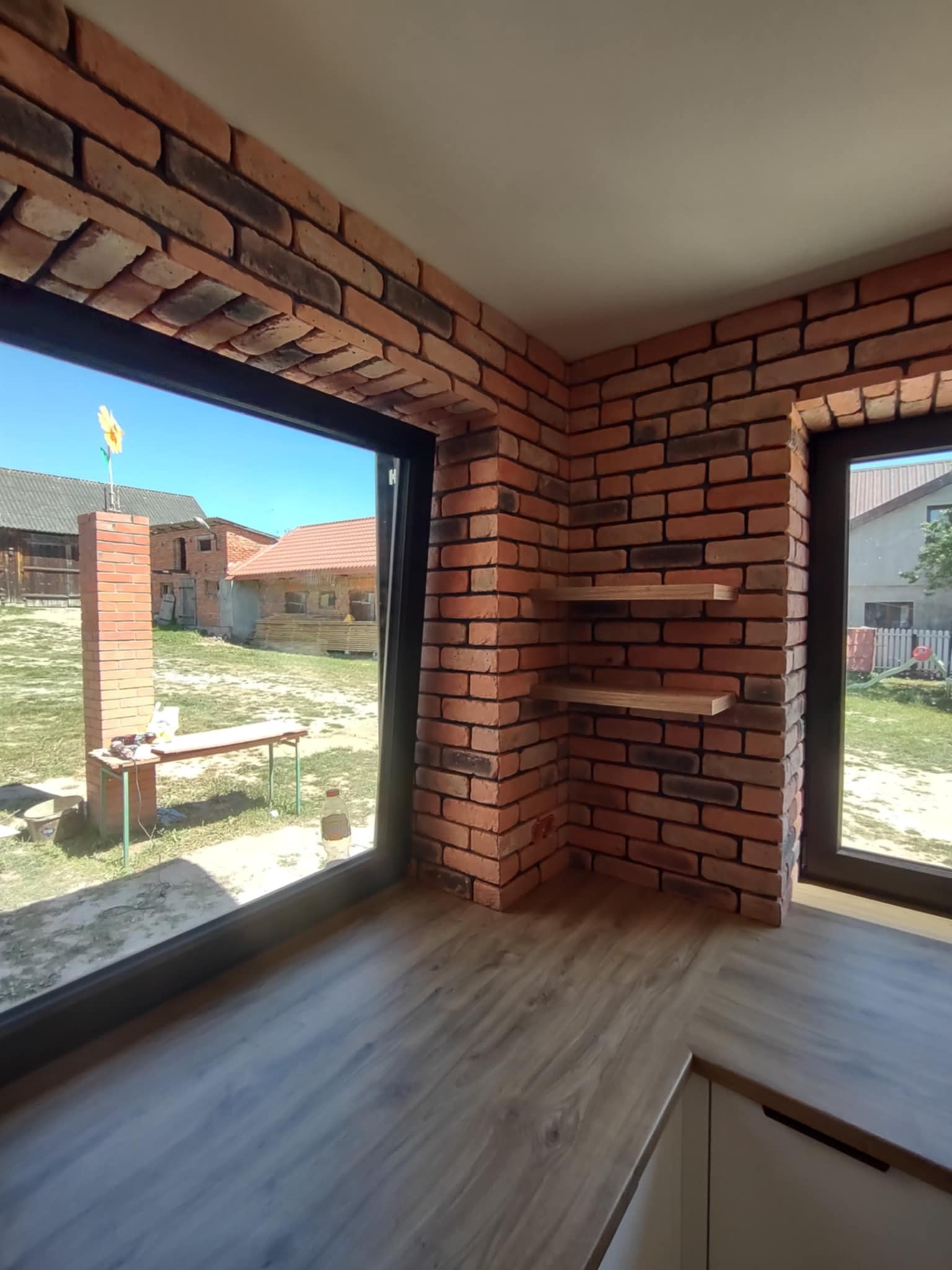 Wnętrze z ceglaną ścianą, drewnianymi półkami i dużym oknem z widokiem na zewnątrz. Podłoga drewniana, blat kuchenny. Perspektywa z narożnika pomieszczenia.