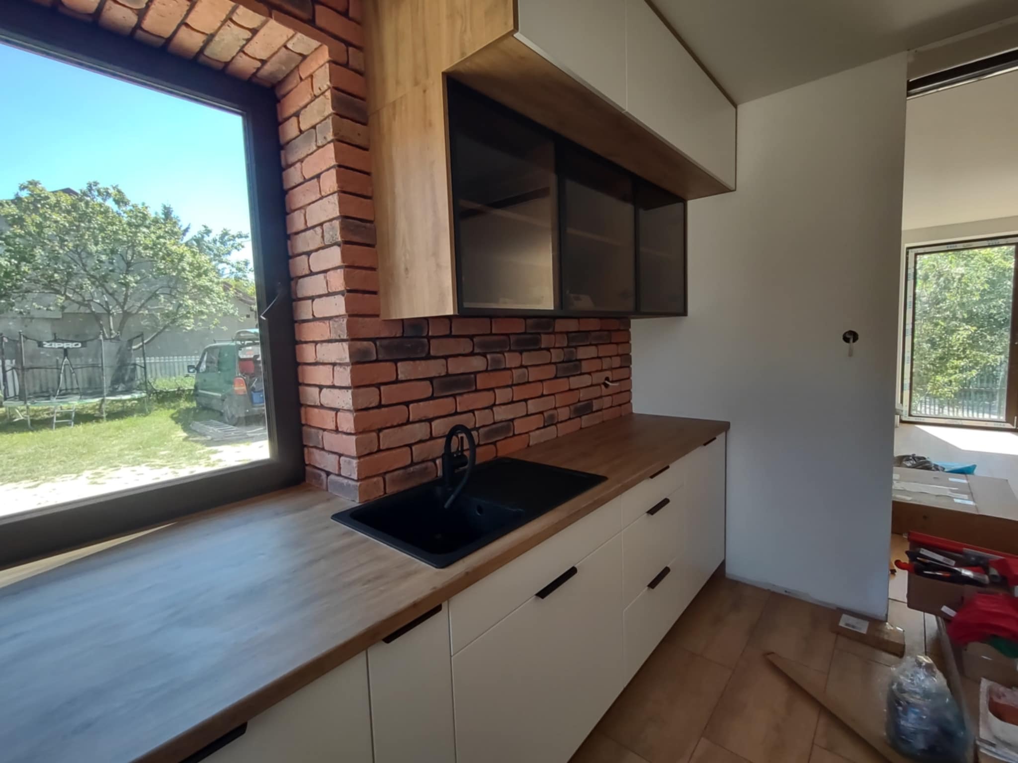 Nowoczesna kuchnia z ceglaną ścianą, drewnianym blatem i czarnym zlewem pod oknem. Białe szafki dolne z czarnymi uchwytami. Częściowo widoczne kartony na podłodze.