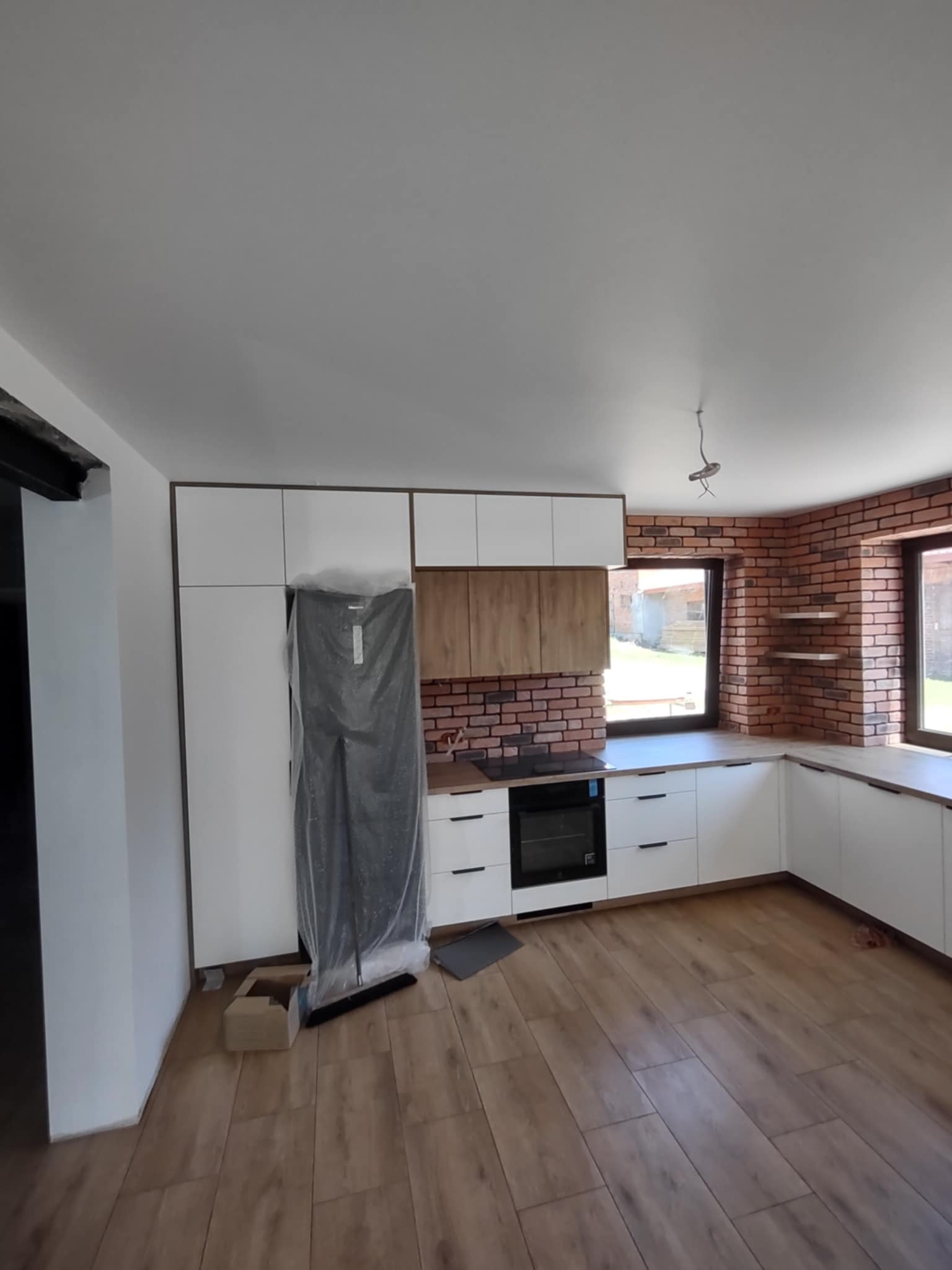 Nowoczesna kuchnia z białymi szafkami, ceglaną ścianą i drewnianą podłogą. Lodówka zabezpieczona folią, widoczne okna i fragment instalacji elektrycznej.