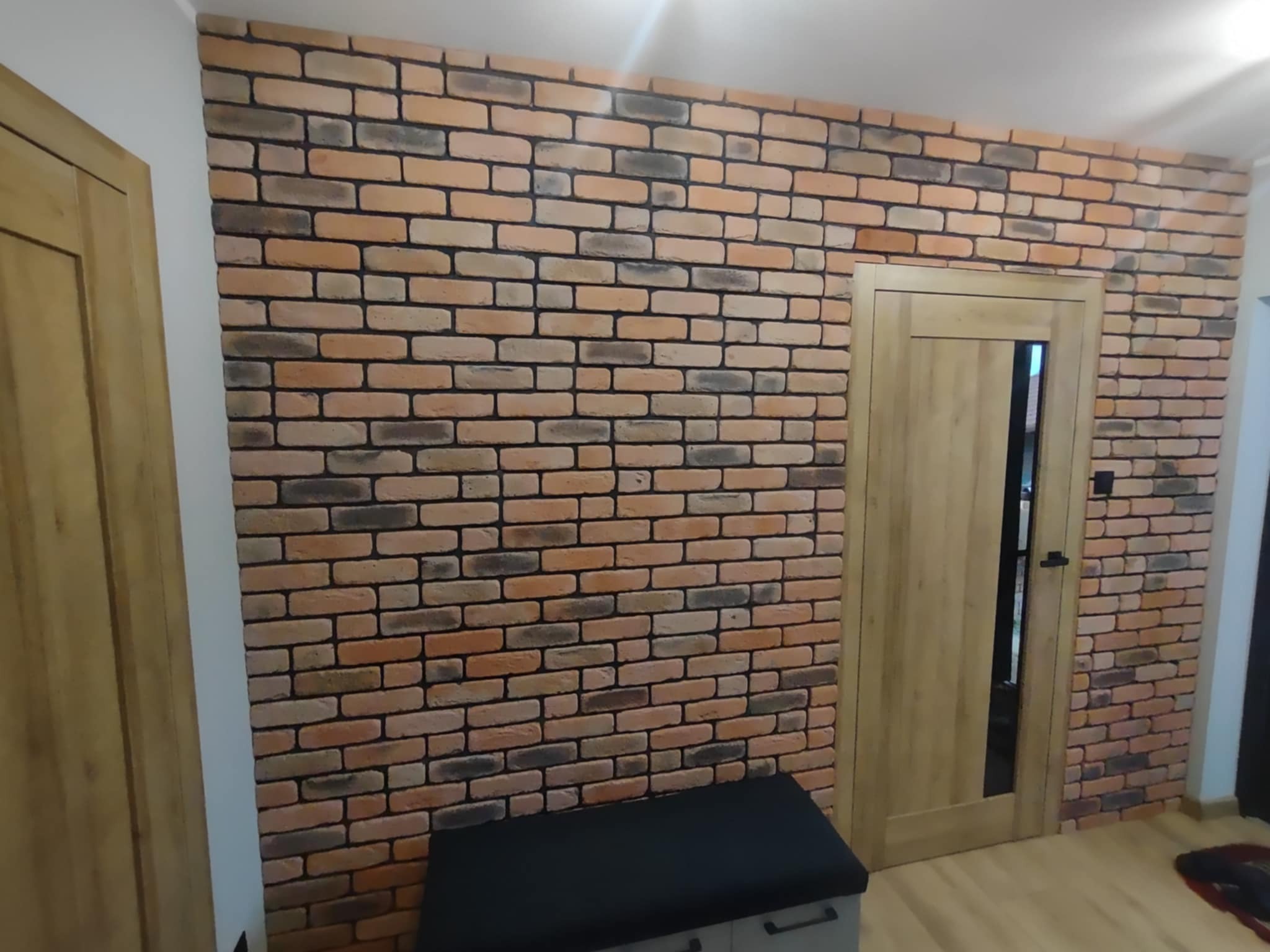 Ściana z cegły klinkierowej w ciepłych odcieniach, z drewnianymi drzwiami i ławką z szufladami. Dekoracyjne wykończenie wnętrza w stylu industrialnym.