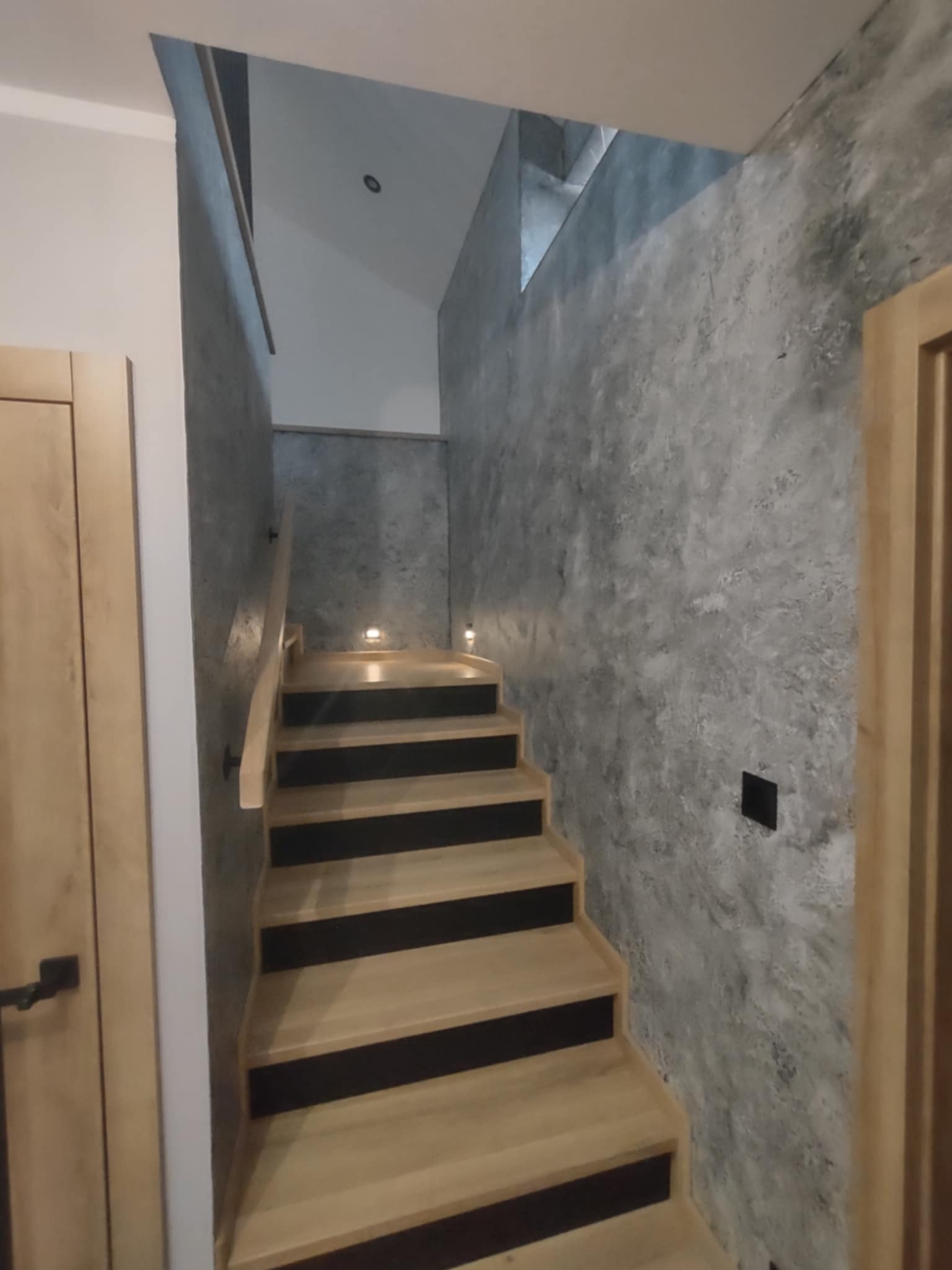 Nowoczesne schody z drewnianymi stopniami i szarą ścianą o fakturze betonu, oświetlone punktowymi lampami. Widok z dołu na górę klatki schodowej.