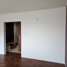 AFINA LUSI SPÓŁKA Z OGRANICZONĄ ODPOWIEDZIALNOŚCIĄ - Czyste, białe ściany z widocznym fragmentem ciemnofioletowej ściany, ciemna drewniana podłoga i otwarta futryna drzwiowa. W tle widać narzędzia i materiały budowlane.