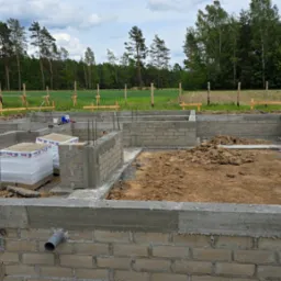 Wzniesione ściany fundamentowe z pustaków betonowych na działce z widocznym zbrojeniem i materiałami budowlanymi. W tle drzewa i polna przestrzeń.