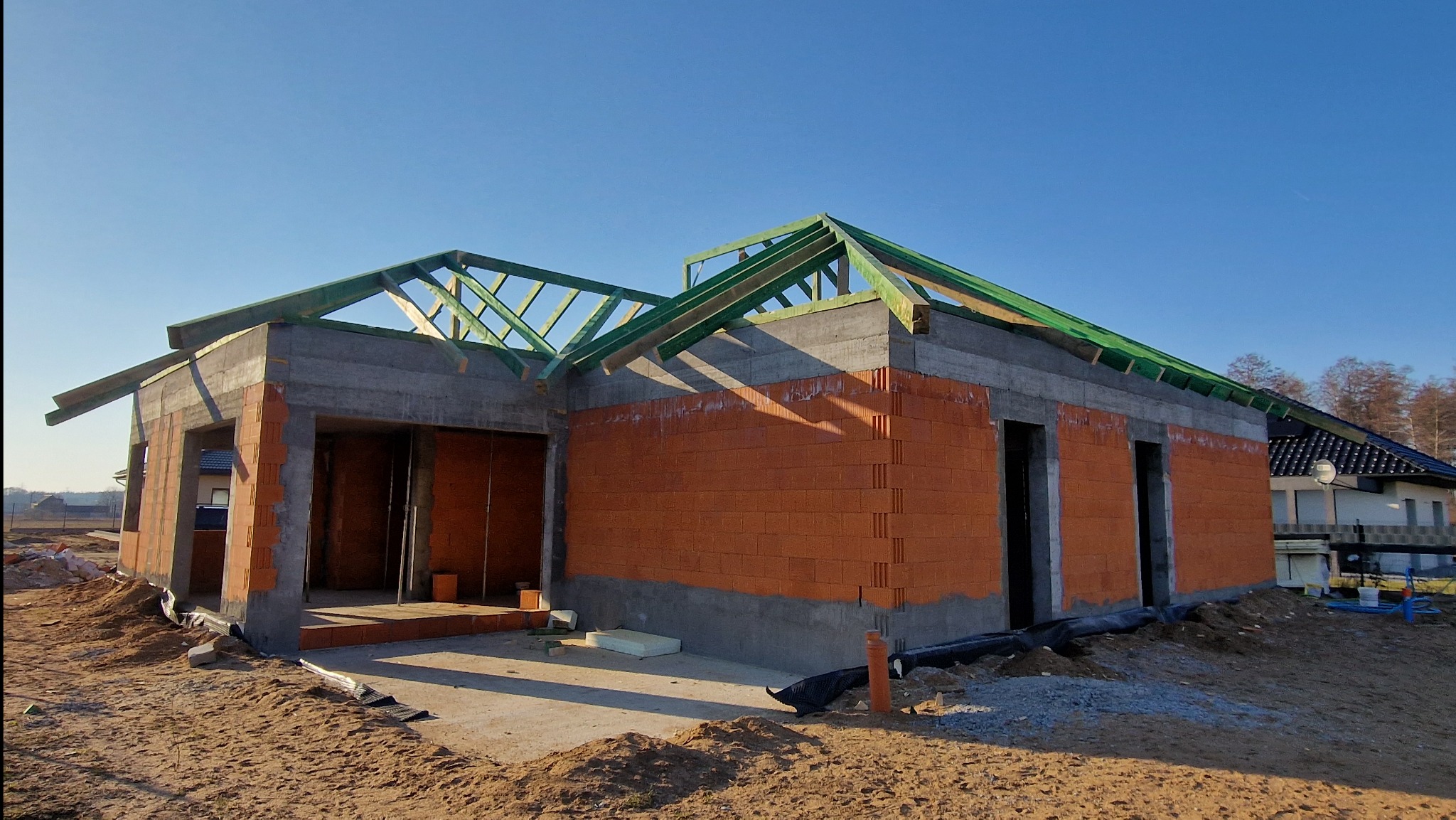 Budowa domu jednorodzinnego: ściany z czerwonej cegły, betonowy wieniec i zielona konstrukcja dachu na tle błękitnego nieba. Widoczne zarysy okien i drzwi.