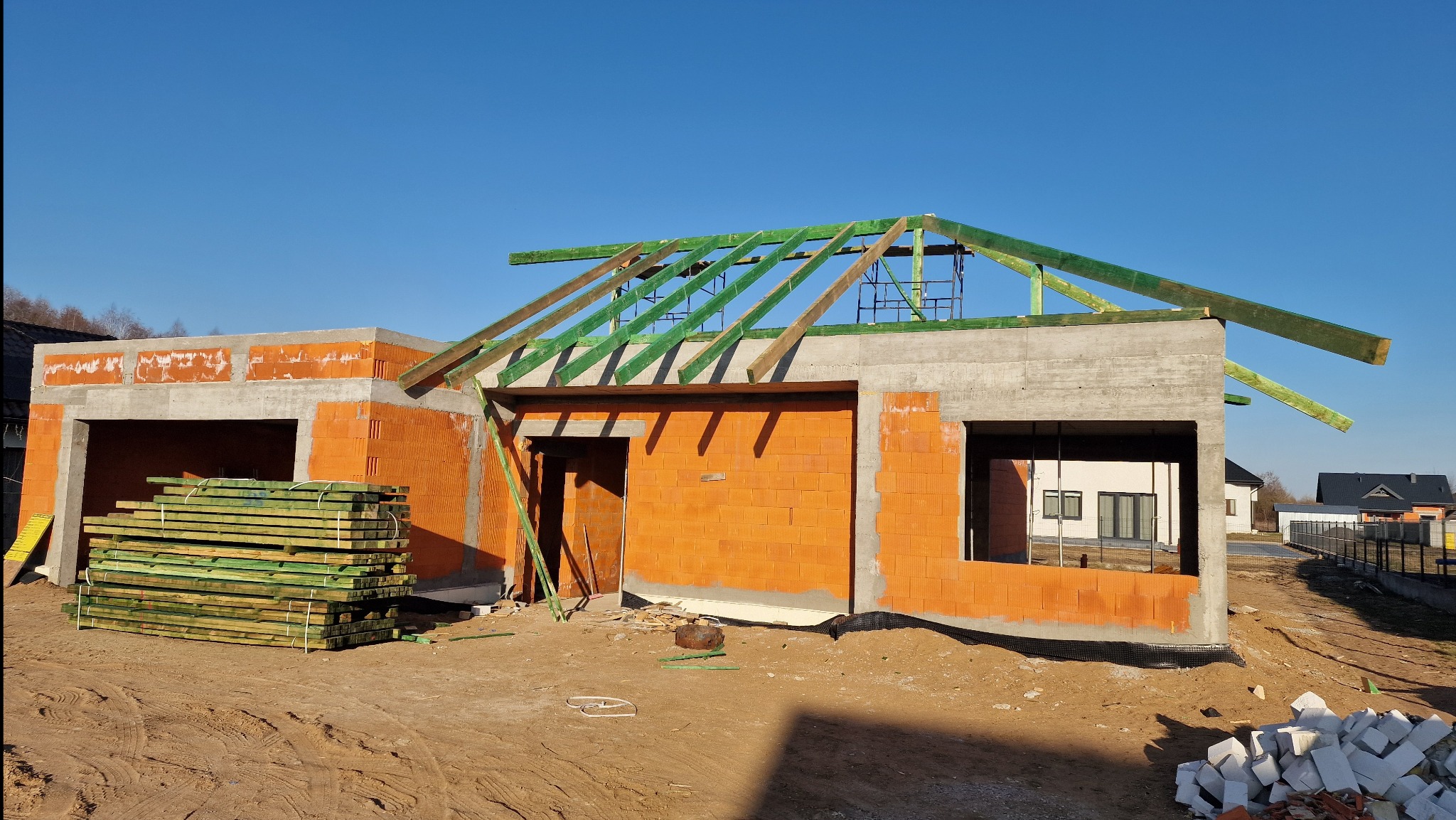 Budowa domu z betonu komórkowego w stanie surowym otwartym, widoczne ściany, strop betonowy i elementy więźby dachowej. Na pierwszym planie paleta z drewnem.