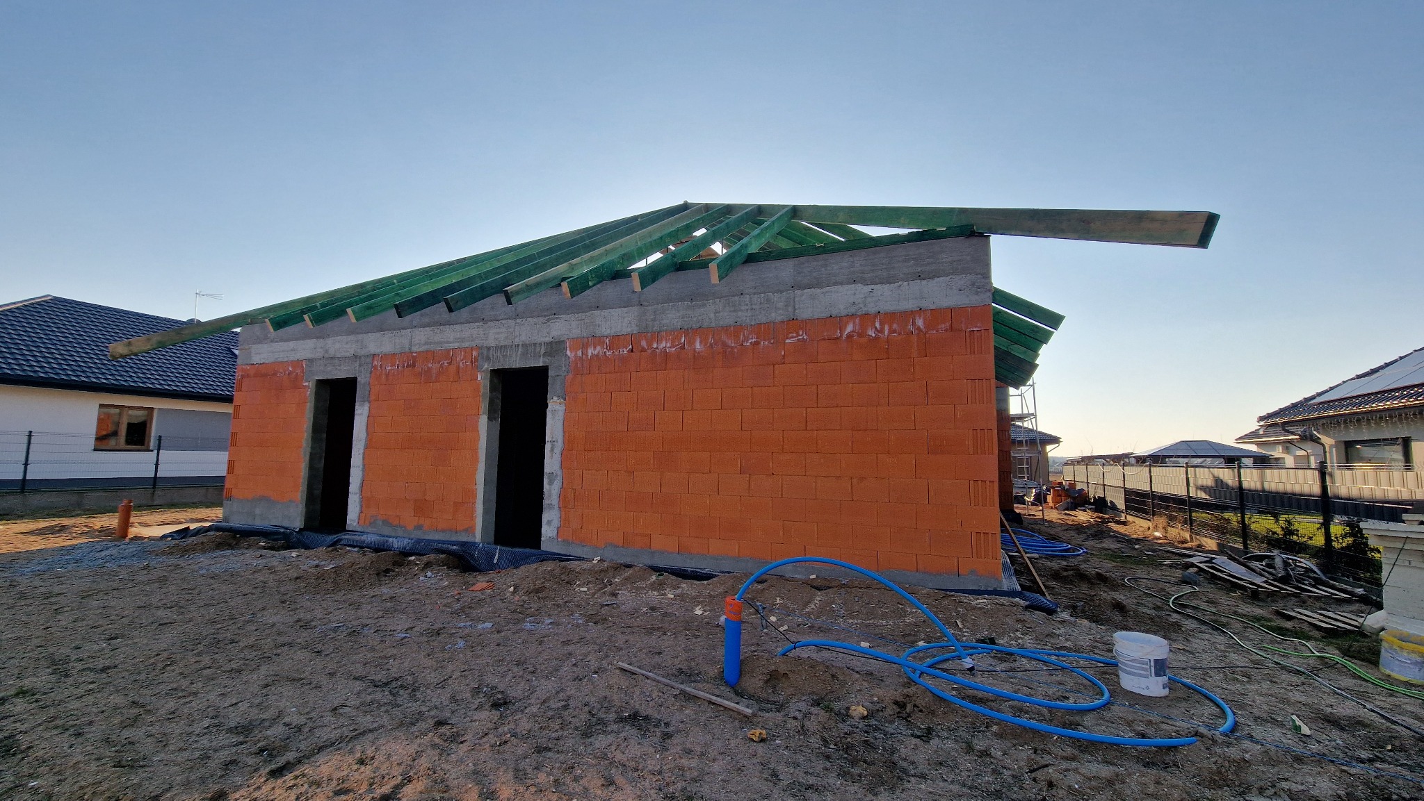 Niski, murowany budynek w stanie surowym otwartym z czerwonej cegły, z widoczną drewnianą konstrukcją dachu i niebieskimi rurami na ziemi, w słoneczny dzień.