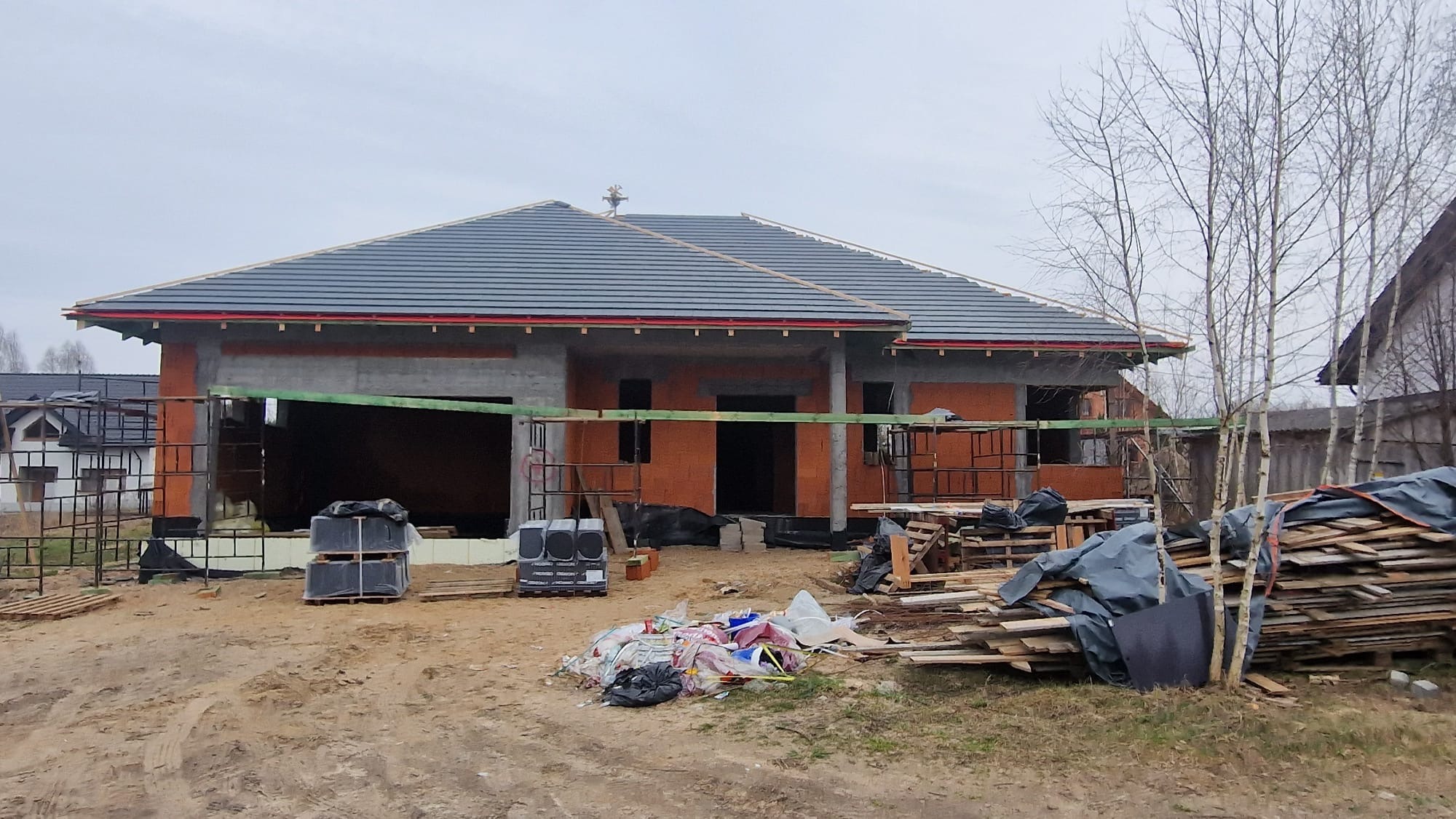 Dom w stanie surowym otwartym z szarą dachówką, widoczne materiały budowlane i rusztowania. Dom z cegły z betonowymi nadprożami i wieńcem.