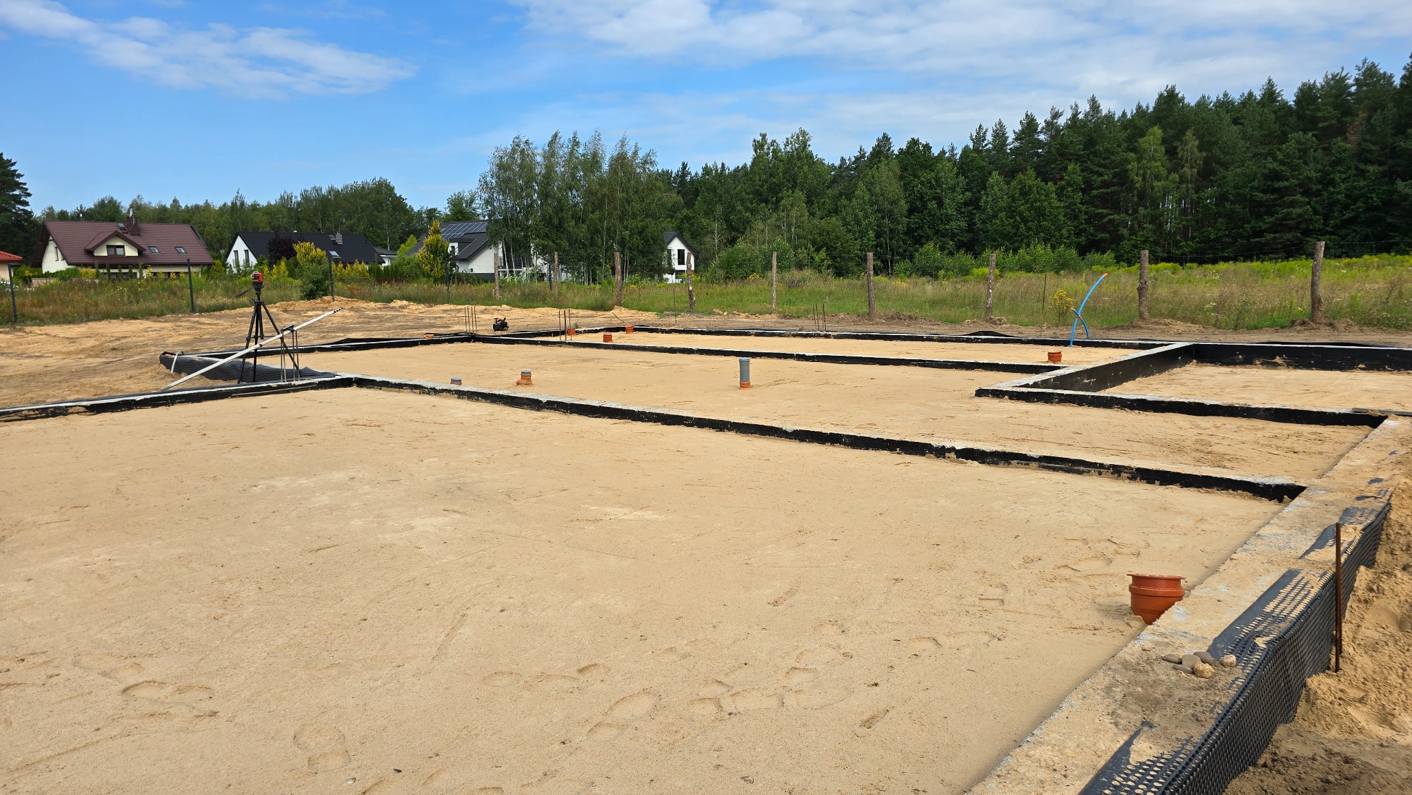 Przygotowany teren pod fundamenty domu z wykopami i izolacją, widoczne rury kanalizacyjne i niwelator laserowy. W tle zabudowa jednorodzinna i las.