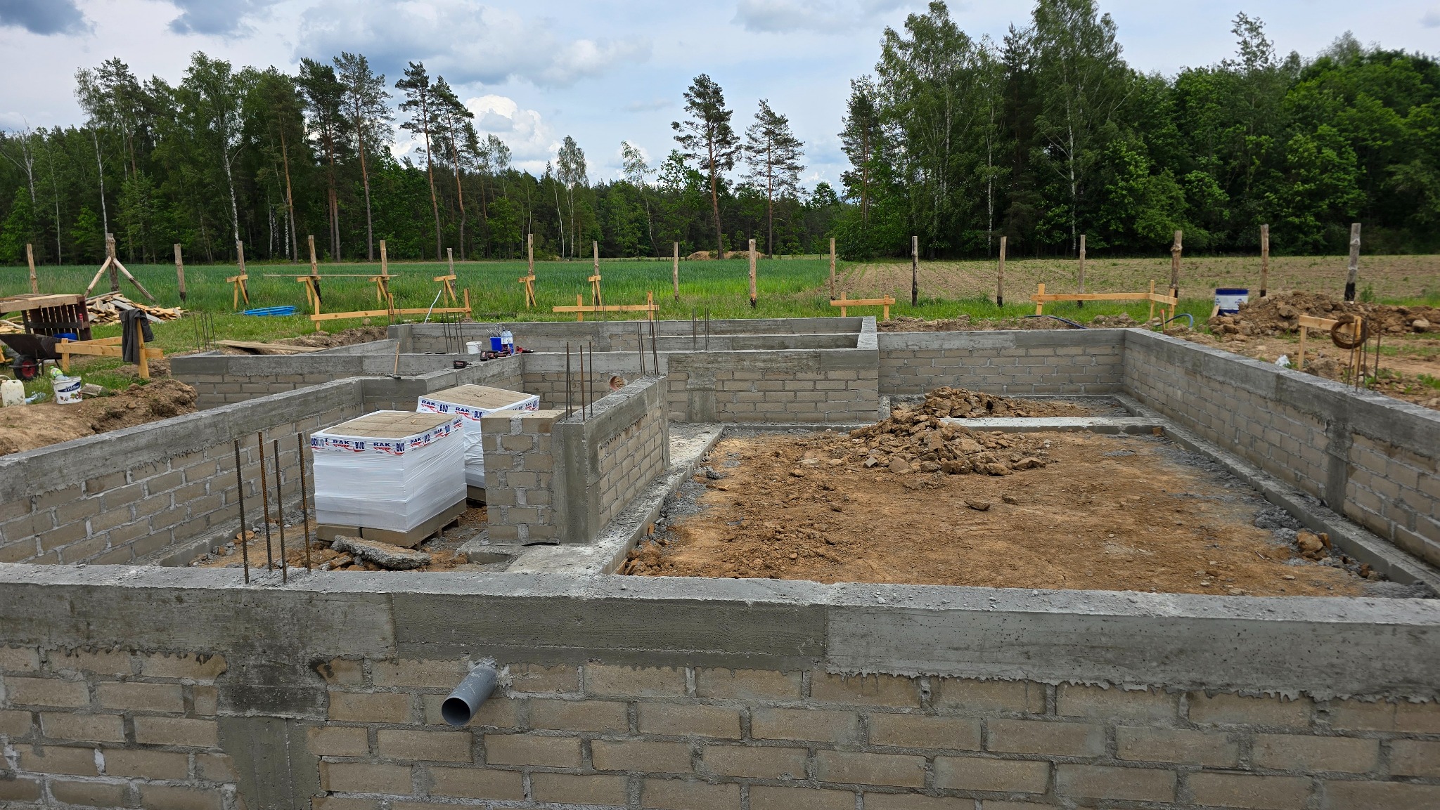 Wzniesione ściany fundamentowe z pustaków betonowych na działce z widocznym zbrojeniem i materiałami budowlanymi. W tle drzewa i polna przestrzeń.