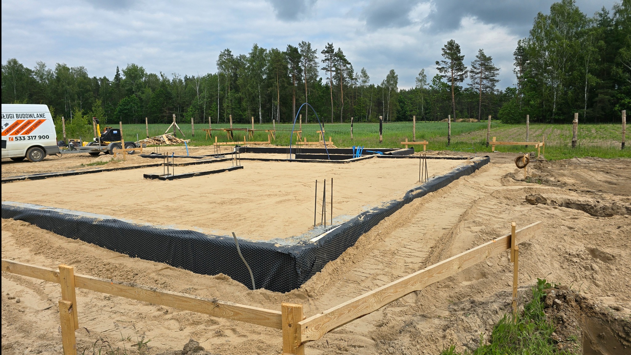 Etap budowy fundamentów: wykop z piaskiem, zbrojenie, izolacja pionowa. Na działce zaparkowany bus firmy budowlanej. W tle widoczny las i pochmurne niebo.