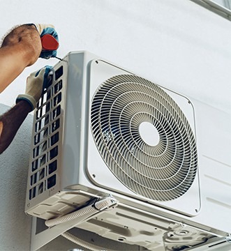 Montaż klimatyzacji: pracownik w rękawicach dokręca śrubę w obudowie jednostki zewnętrznej klimatyzatora na jasnej ścianie. Widoczne przewody.