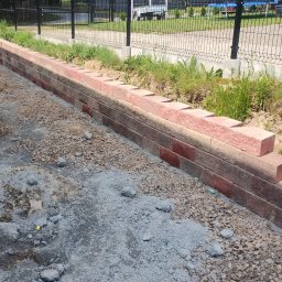 Robótka Na Luzie - Świeżo zbudowany murek oporowy z czerwonej cegły, widoczny na tle zieleni i ogrodzenia. Ułożone bloki tworzą solidną konstrukcję.