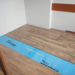 Układanie paneli podłogowych w jasnym wnętrzu. Część podłogi wyłożona jest niebieską folią izolacyjną. Widoczny fragment poziomicy.