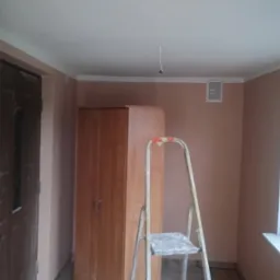 Pokój po odświeżeniu: ściany w odcieniach beżu, biały sufit i listwy. Widoczna drabina malarska, drewniana szafa i fragment okna. Kabel zwisający z sufitu.