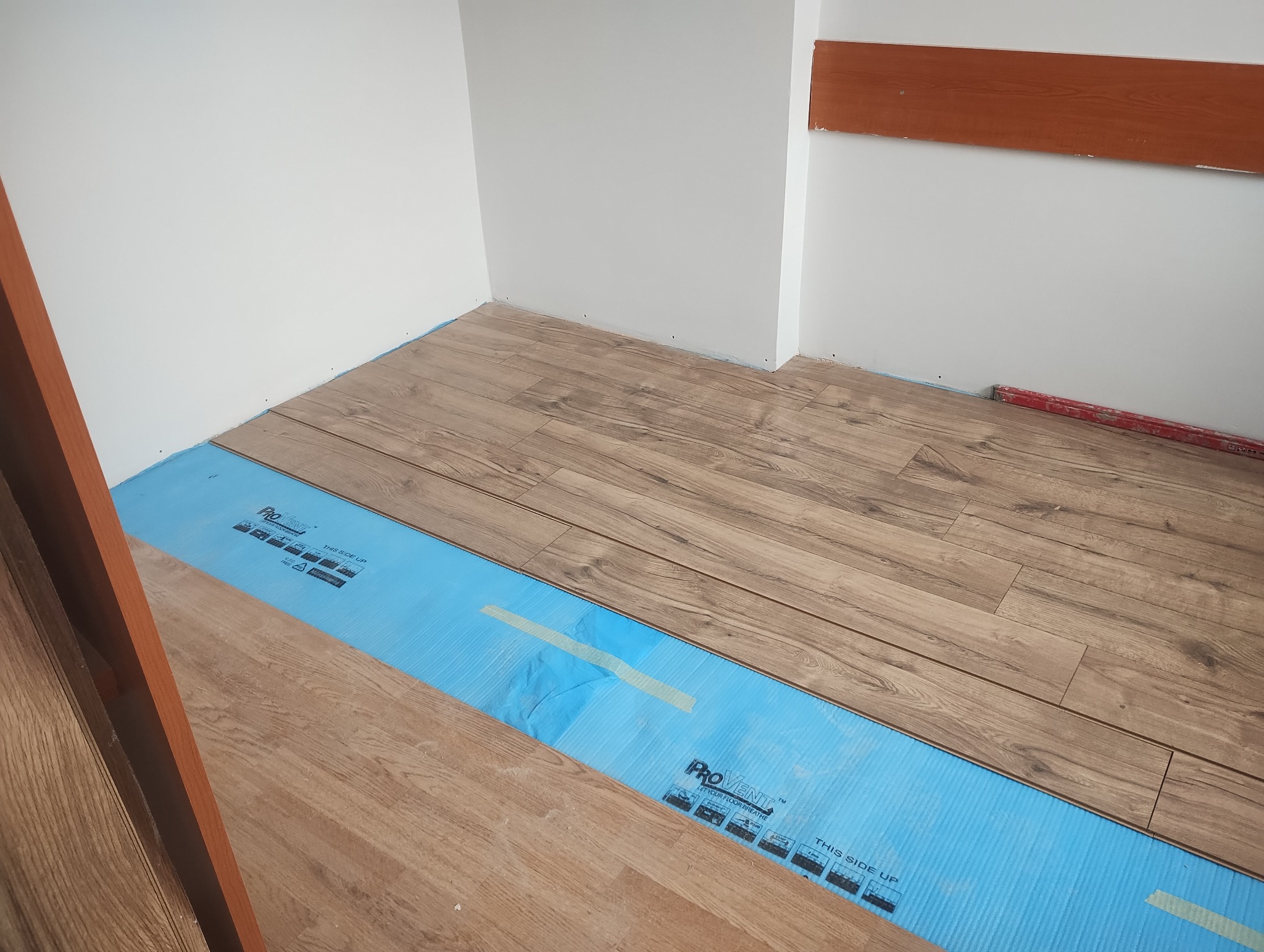 Układanie paneli podłogowych w jasnym wnętrzu. Część podłogi wyłożona jest niebieską folią izolacyjną. Widoczny fragment poziomicy.