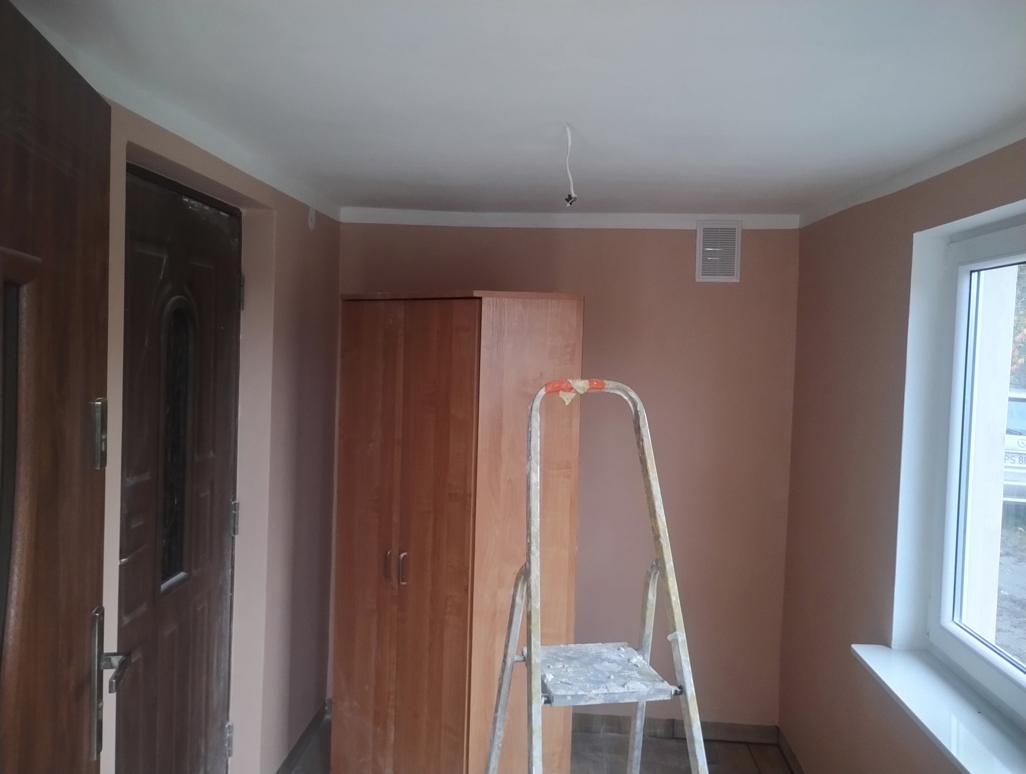 Pokój po odświeżeniu: ściany w odcieniach beżu, biały sufit i listwy. Widoczna drabina malarska, drewniana szafa i fragment okna. Kabel zwisający z sufitu.