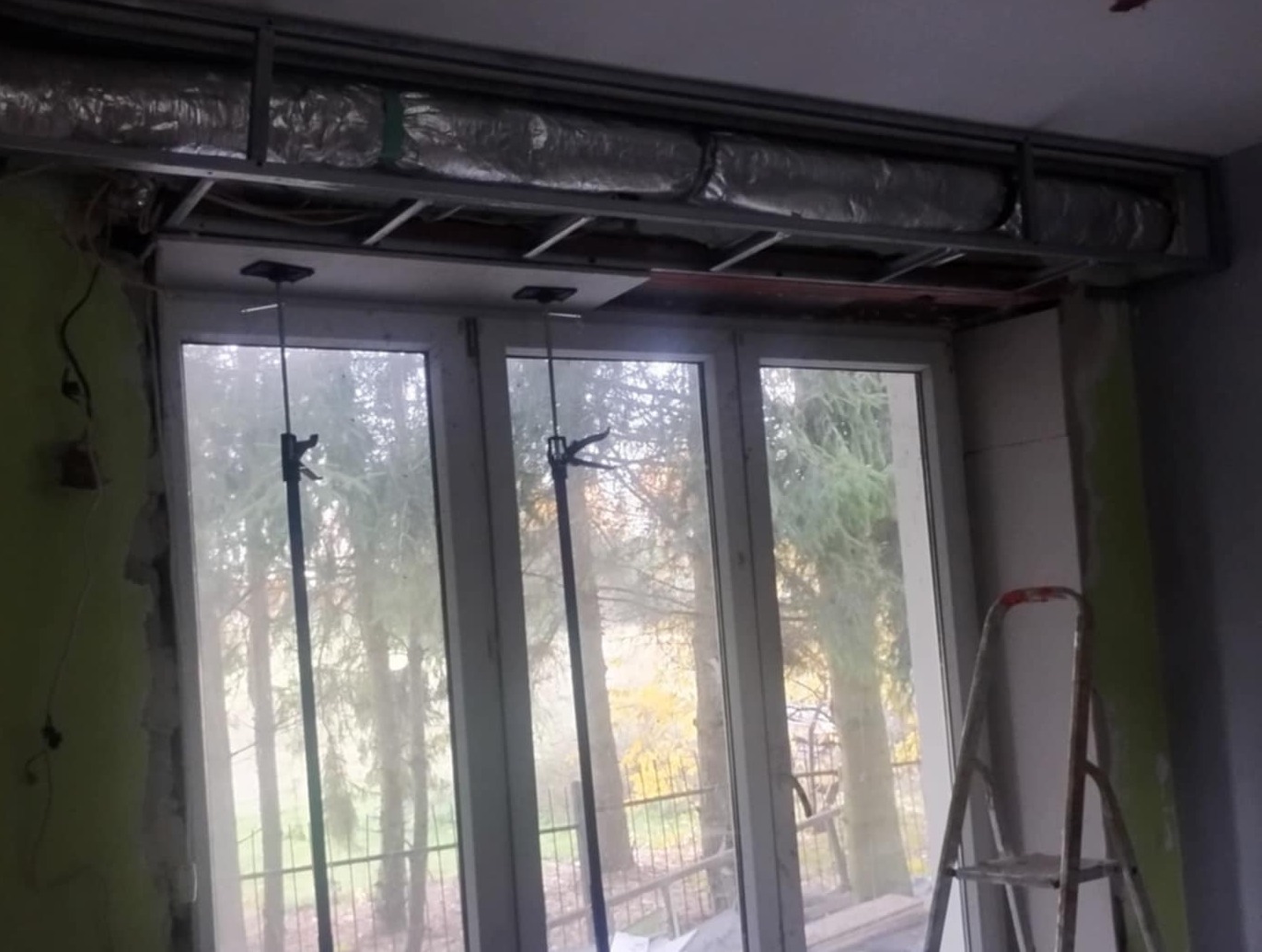 Montaż sufitu podwieszanego z płyt karton-gips w pomieszczeniu z oknem. Widoczne profile, izolacja i drabina. Etap prac przed wykończeniem.