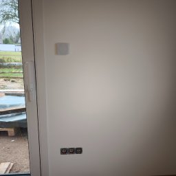 Adrian Duda - Usługi elektryczne - Biała ściana z trzema gniazdkami elektrycznymi bez osprzętu, obok okno z widokiem na zewnątrz i panel sterowania na ścianie. Etap prac wykończeniowych.