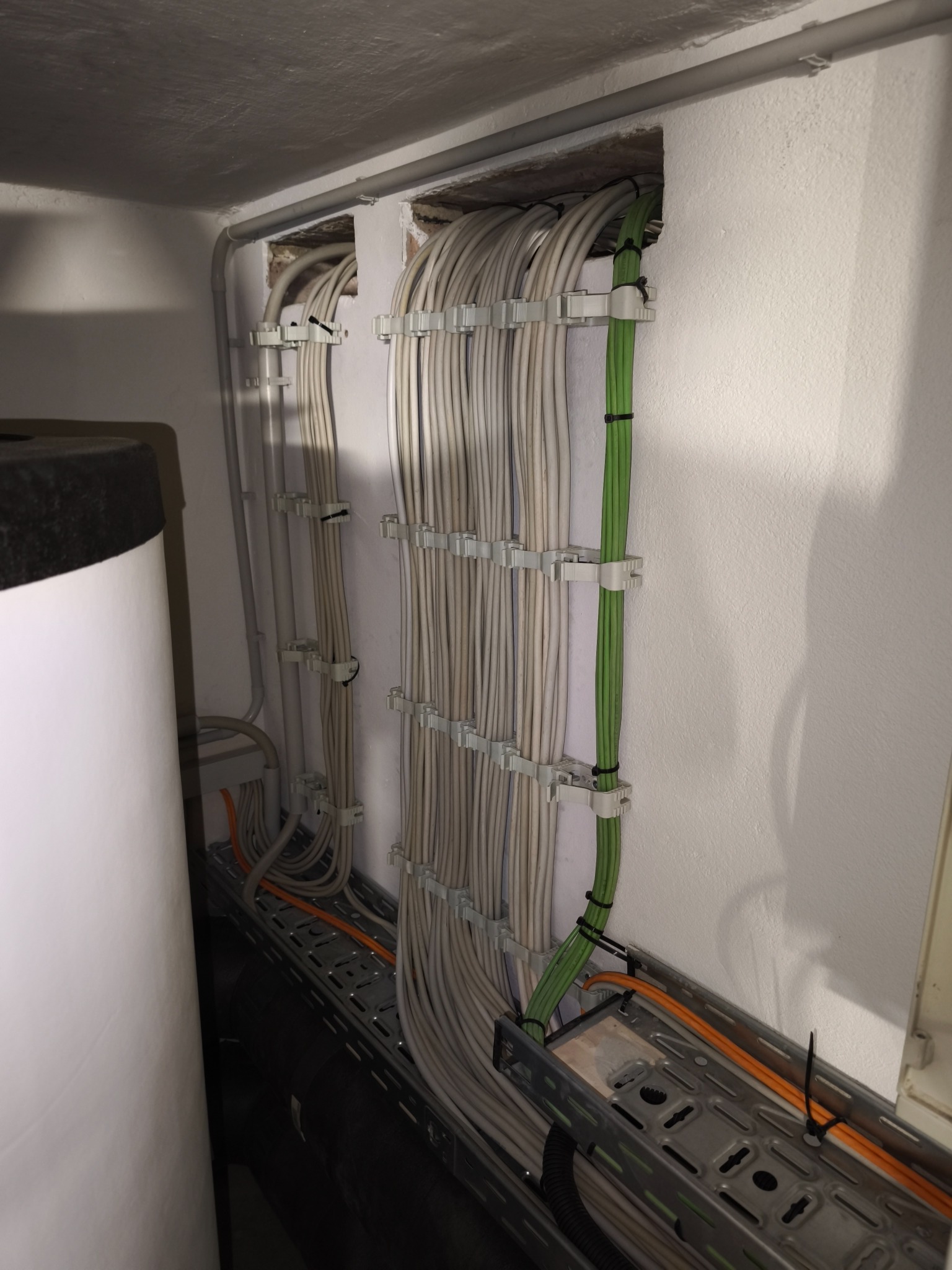 Równolegle ułożone kable elektryczne w korytkach, przymocowane do ściany za pomocą uchwytów. Widoczne kable szare i zielone, oraz fragment bojlera.