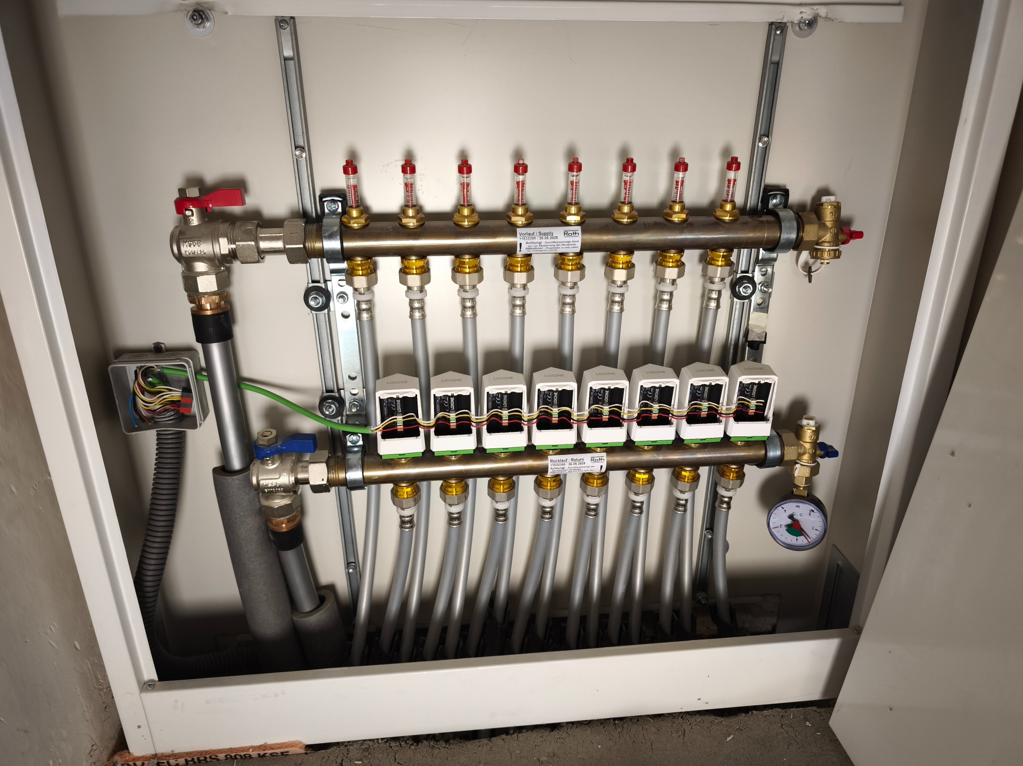 Rozdzielacz ogrzewania podłogowego w szafce, z zaworami, przepływomierzami i przewodami. Widoczna instalacja elektryczna sterująca systemem.