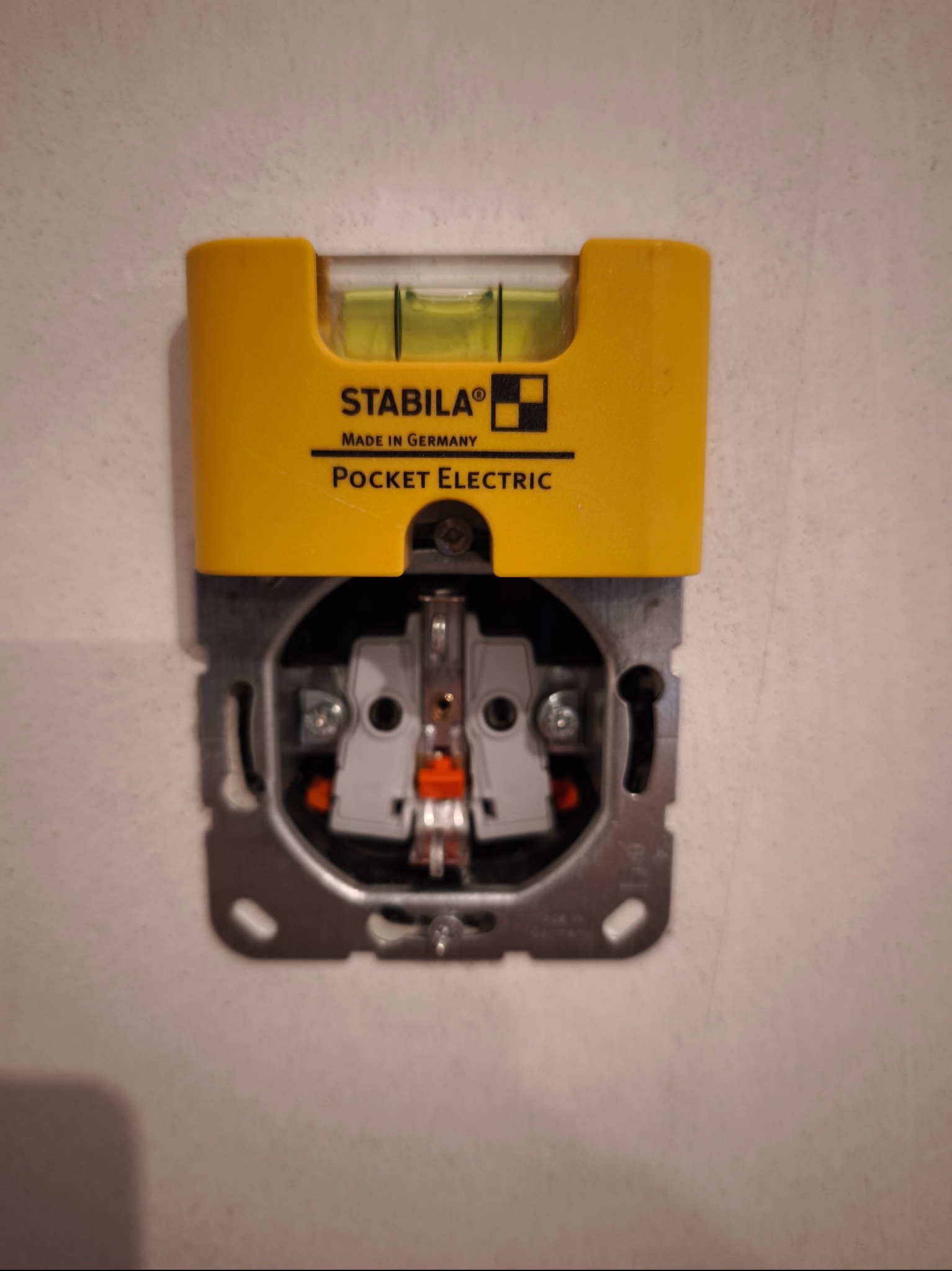 Żółta poziomica Stabila Pocket Electric na metalowej ramce gniazdka elektrycznego, zamontowanego na białej ścianie. Widoczne elementy wewnętrzne gniazdka.