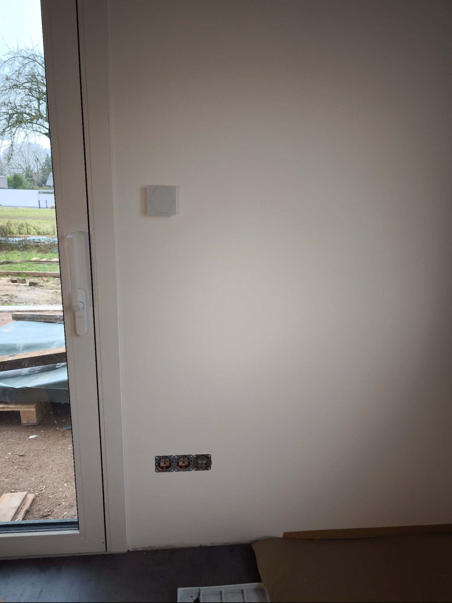 Biała ściana z trzema gniazdkami elektrycznymi bez osprzętu, obok okno z widokiem na zewnątrz i panel sterowania na ścianie. Etap prac wykończeniowych.