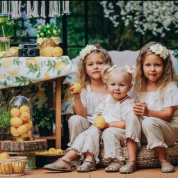 Weronika Kunek - Trzy urocze dziewczynki z blond włosami, w białych ubraniach, siedzą na tle dekoracji z motywem cytrynowym. Dziewczynki trzymają cytryny.