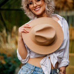 Weronika Kunek - Uśmiechnięta blondynka w okularach pozuje z beżowym kapeluszem, ubrana w białą koszulę i dżinsy z ozdobnym paskiem. Naturalne światło, plener.