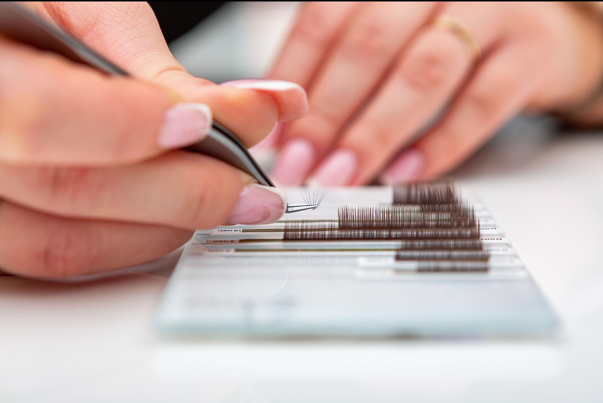 Stylistka rzęs precyzyjnie aplikuje kępkę sztucznych rzęs pęsetą, zbliżenie na dłonie i materiały, delikatny manicure.