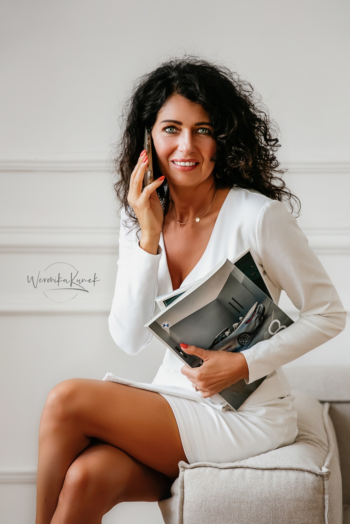 Kobieta w białej sukience siedzi, rozmawia przez telefon i trzyma magazyn z samochodem BMW, uśmiechając się do kamery. Sesja wizerunkowa.