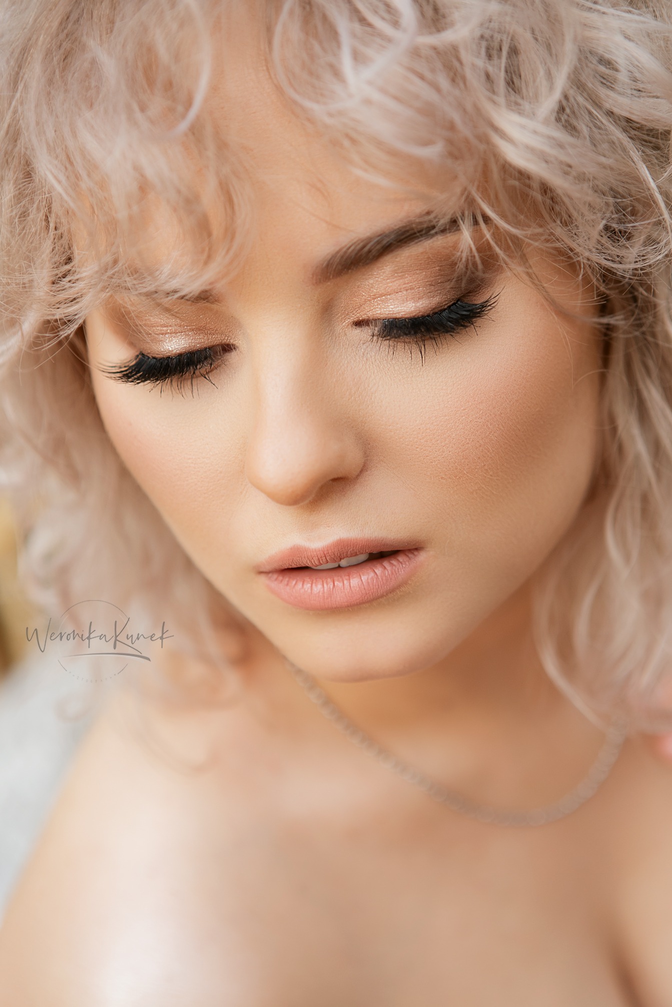 Portret kobiety z blond kręconymi włosami, delikatnym makijażem i srebrnym naszyjnikiem. Zamknięte oczy, skupienie na detalu urody. Naturalne światło.