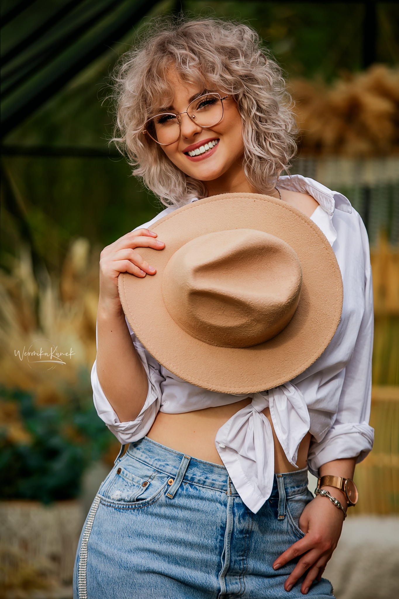 Uśmiechnięta blondynka w okularach pozuje z beżowym kapeluszem, ubrana w białą koszulę i dżinsy z ozdobnym paskiem. Naturalne światło, plener.