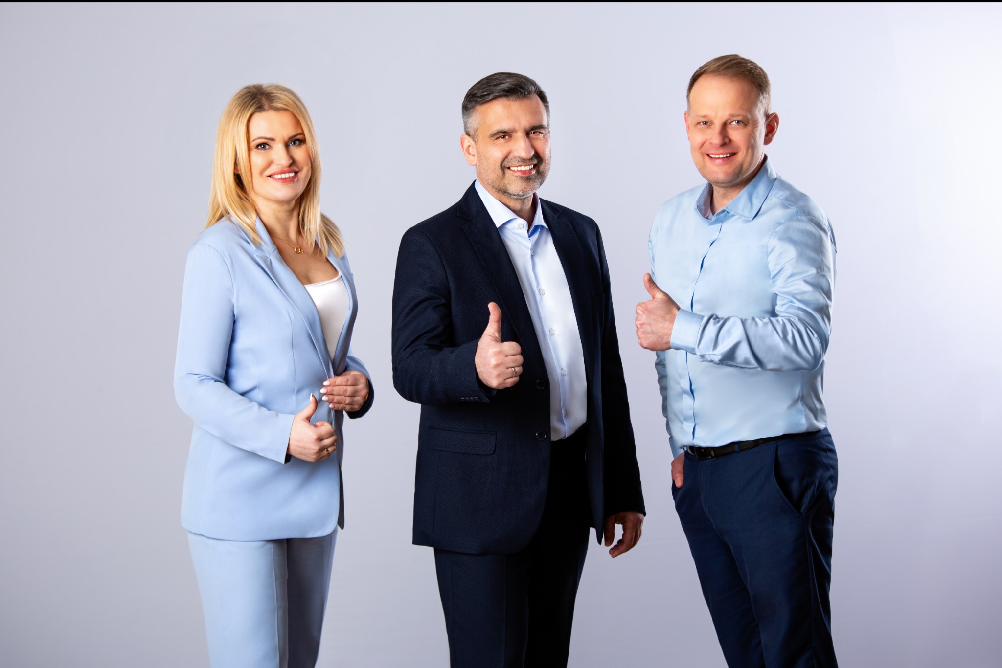 Profesjonalna sesja wizerunkowa: troje uśmiechniętych osób w eleganckich strojach, gestykulujących aprobatę na jasnym tle.