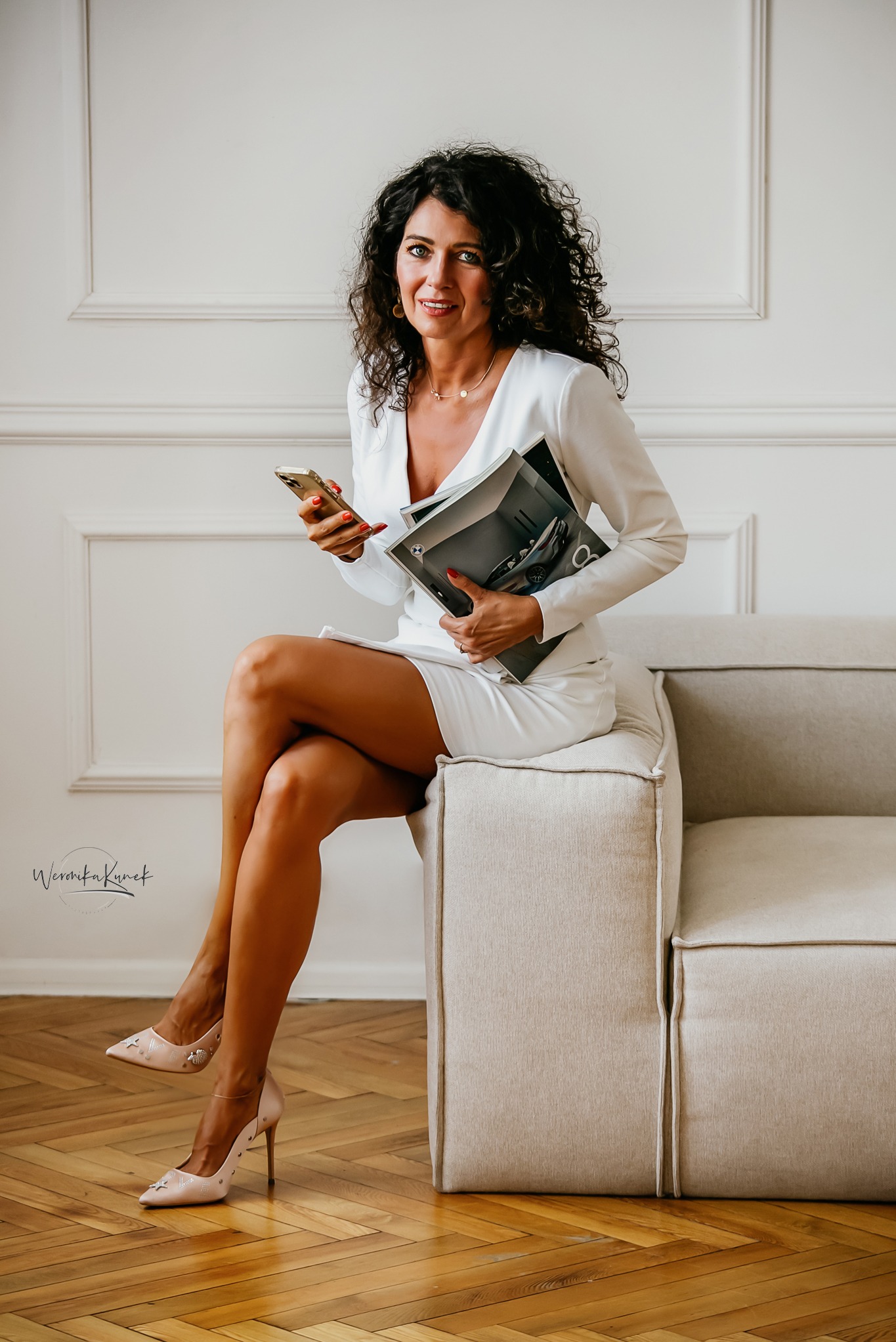 Kobieta w białym garniturze siedzi na sofie z telefonem i magazynem. Elegancka sesja wizerunkowa z subtelnym uśmiechem i naturalnym światłem.