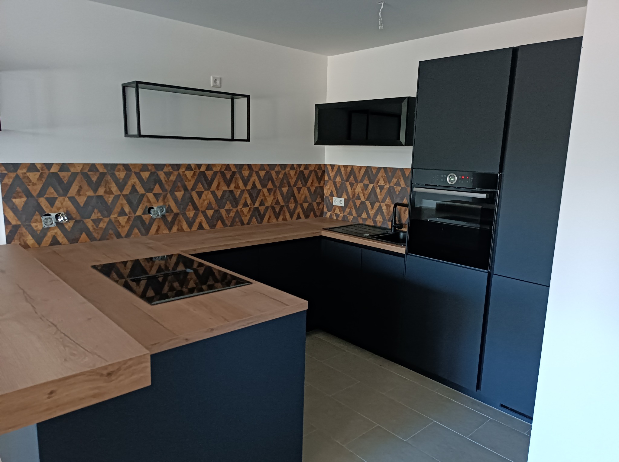 Nowoczesna kuchnia w odcieniach granatu i drewna. Geometryczny wzór na ścianie. Czarna zabudowa z piekarnikiem i płytą grzewczą. Minimalistyczna półka na ścianie.