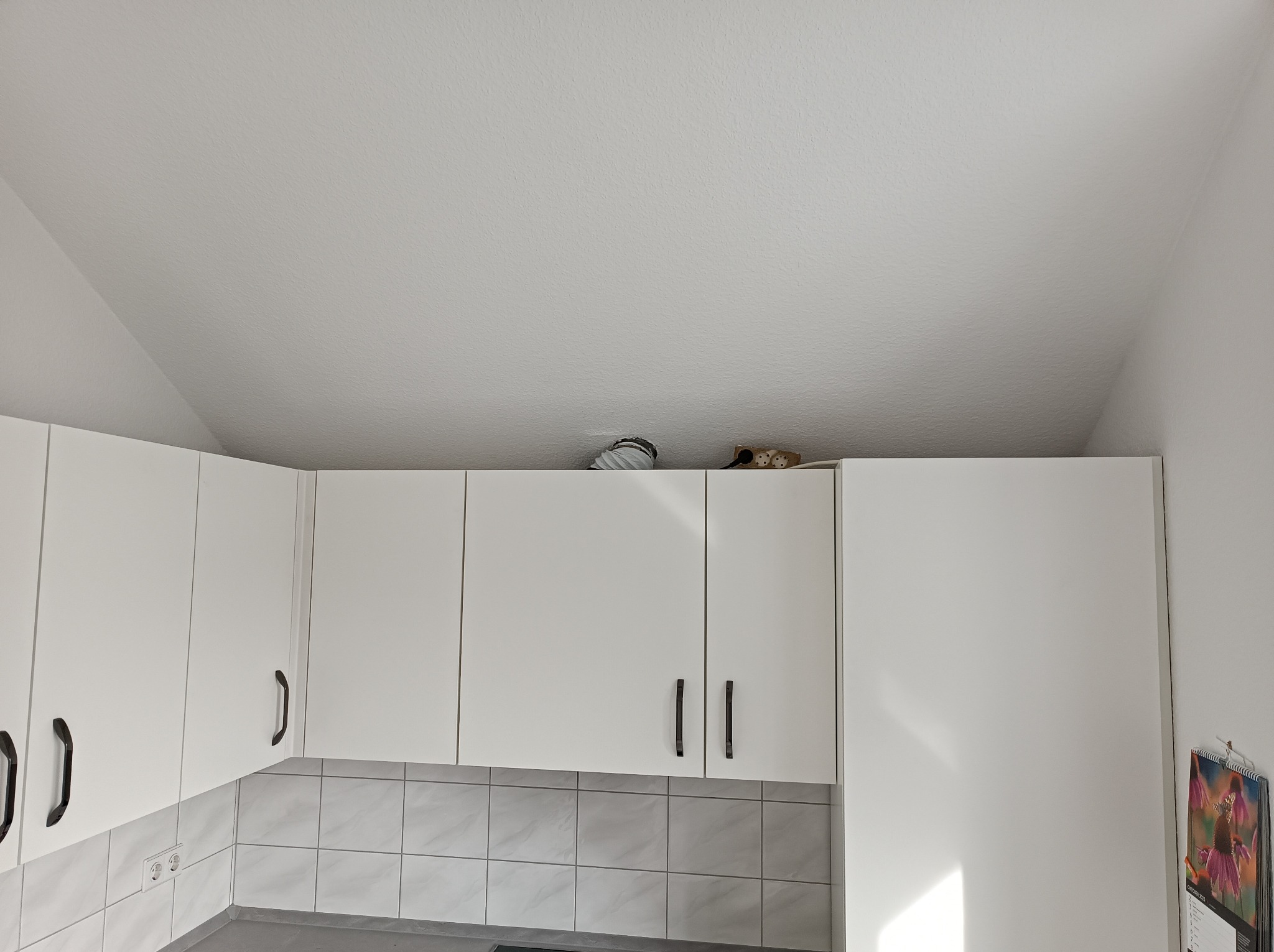 Białe, górne szafki kuchenne z czarnymi uchwytami na tle szarych płytek. Na szafkach widoczne elementy instalacji i akcesoria kuchenne.