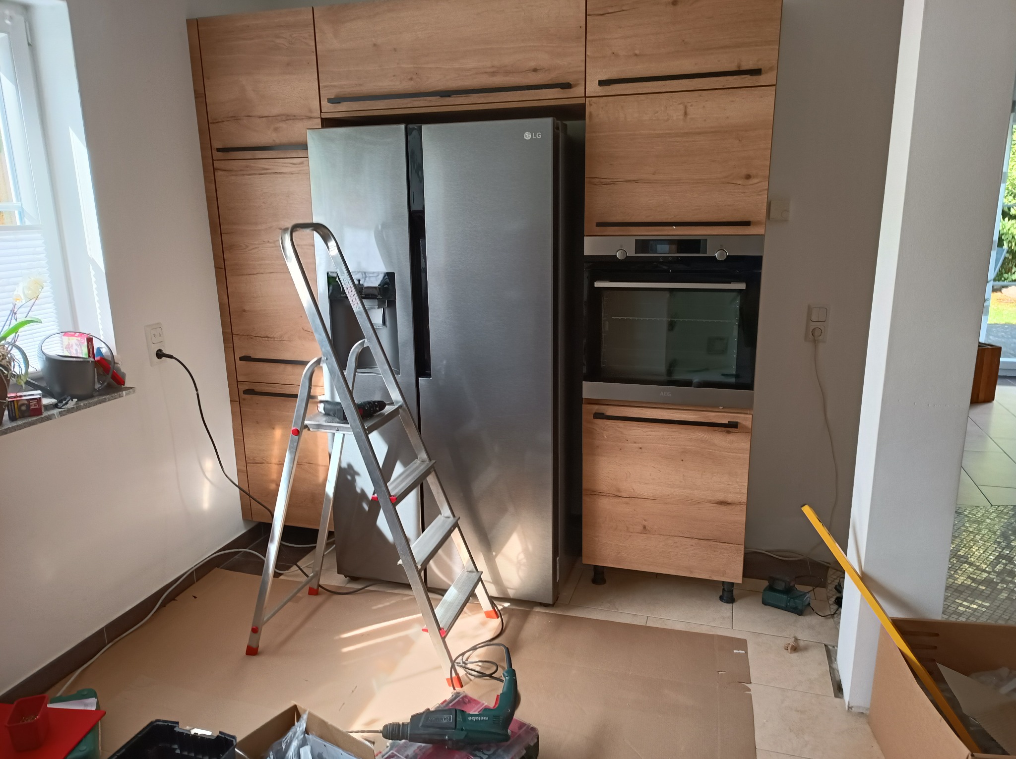 Kuchnia w trakcie montażu: drewniane szafki, srebrna lodówka LG, drabina i wiertarka Metabo na kartonowym podłożu. Widoczny piekarnik i okno.