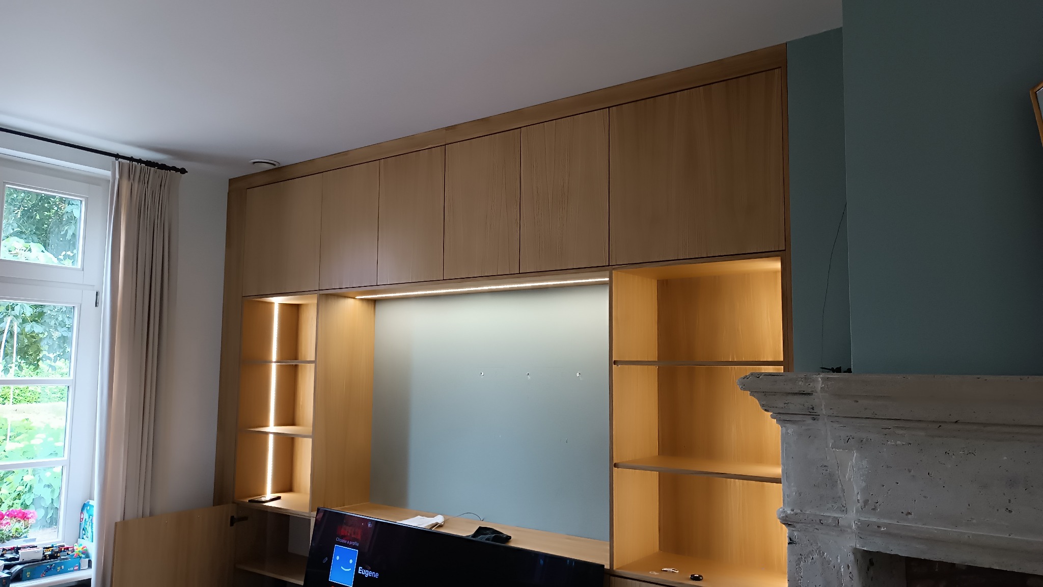 Jasne, drewniane meble na wymiar z podświetlanymi półkami w salonie, widok na okno z zasłonami po lewej, dekoracyjny kominek po prawej stronie.