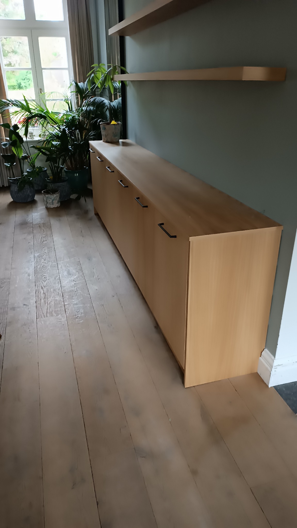 Długi, jasny regał z czarnymi uchwytami w nowoczesnym wnętrzu z drewnianą podłogą i roślinami doniczkowymi przy oknie. Nad regałem widoczne półki.