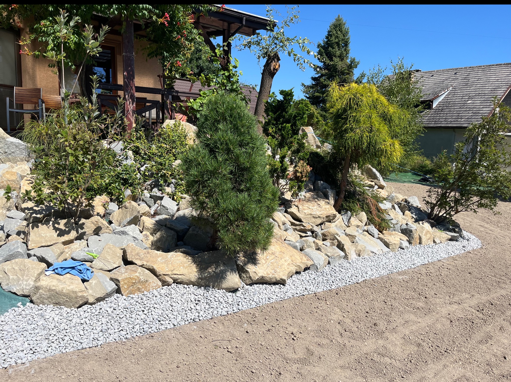 Ogród skalny z nasadzeniami i obrzeżem z drobnych kamieni. Na pierwszym planie przygotowany teren pod trawnik. W tle widoczny dom z elementami drewnianymi.
