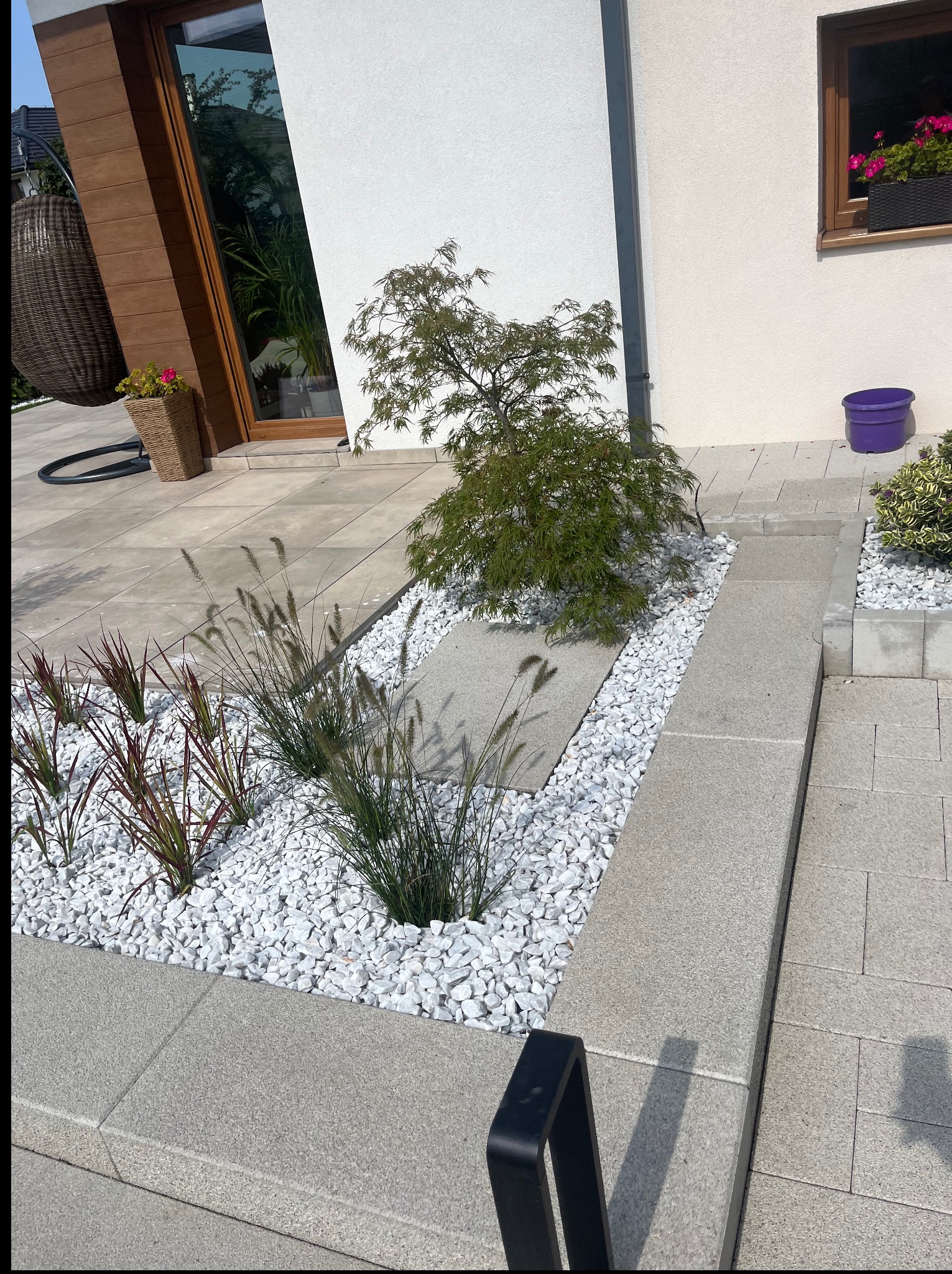 Nowoczesny ogród przydomowy z białymi kamieniami, ozdobnymi trawami i małym drzewkiem japońskim na tle jasnej elewacji domu z drewnianymi elementami.