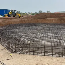 Zbrojenie fundamentu w formie siatki na betonowej płycie, w tle koparka i kontener na budowie. Widok na plac budowy w pochmurny dzień.