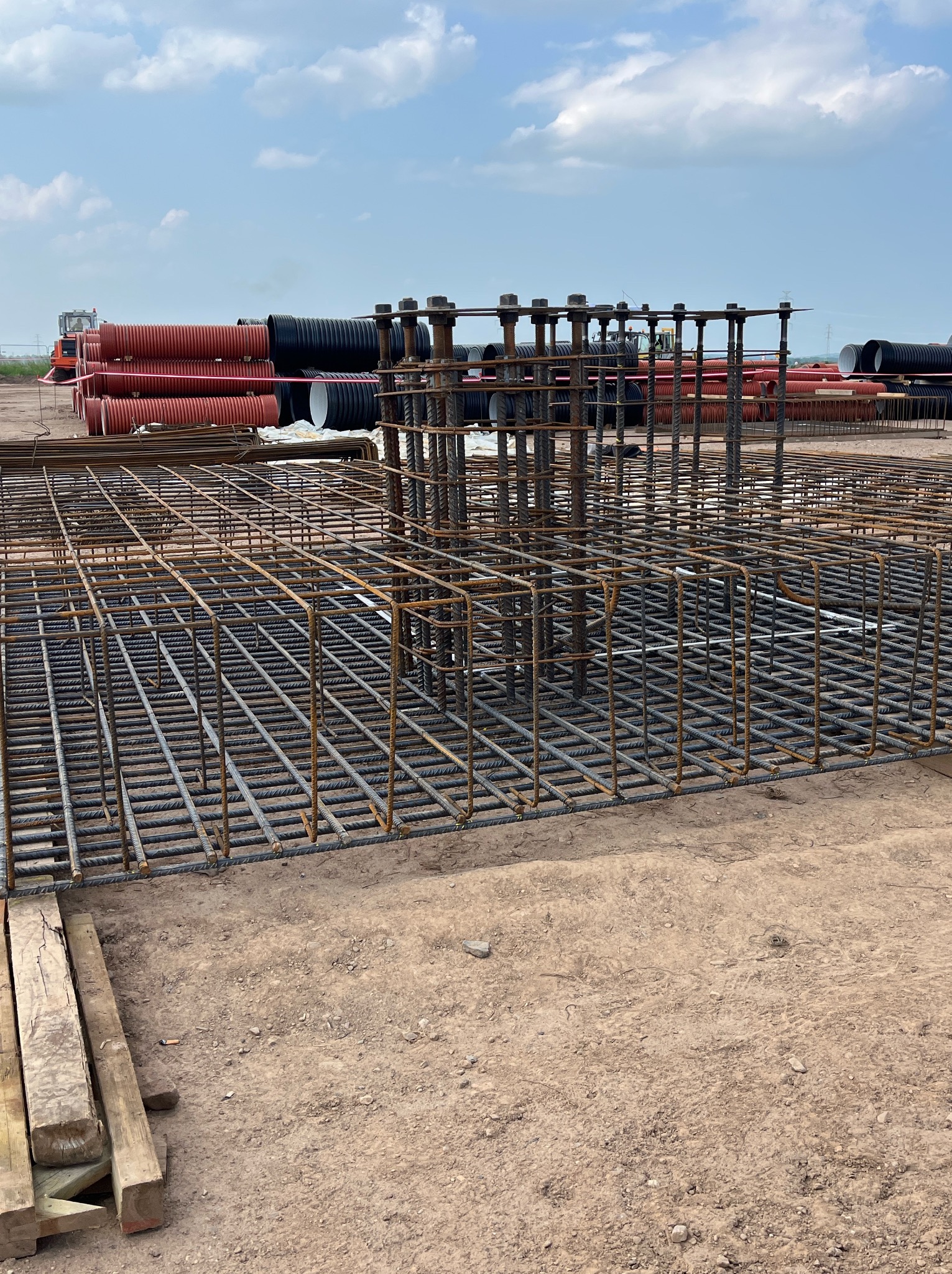 Siatka zbrojeniowa fundamentu z widocznymi prętami i szpilkami, rury drenażowe w tle, teren budowy. Detal konstrukcji żelbetowej.