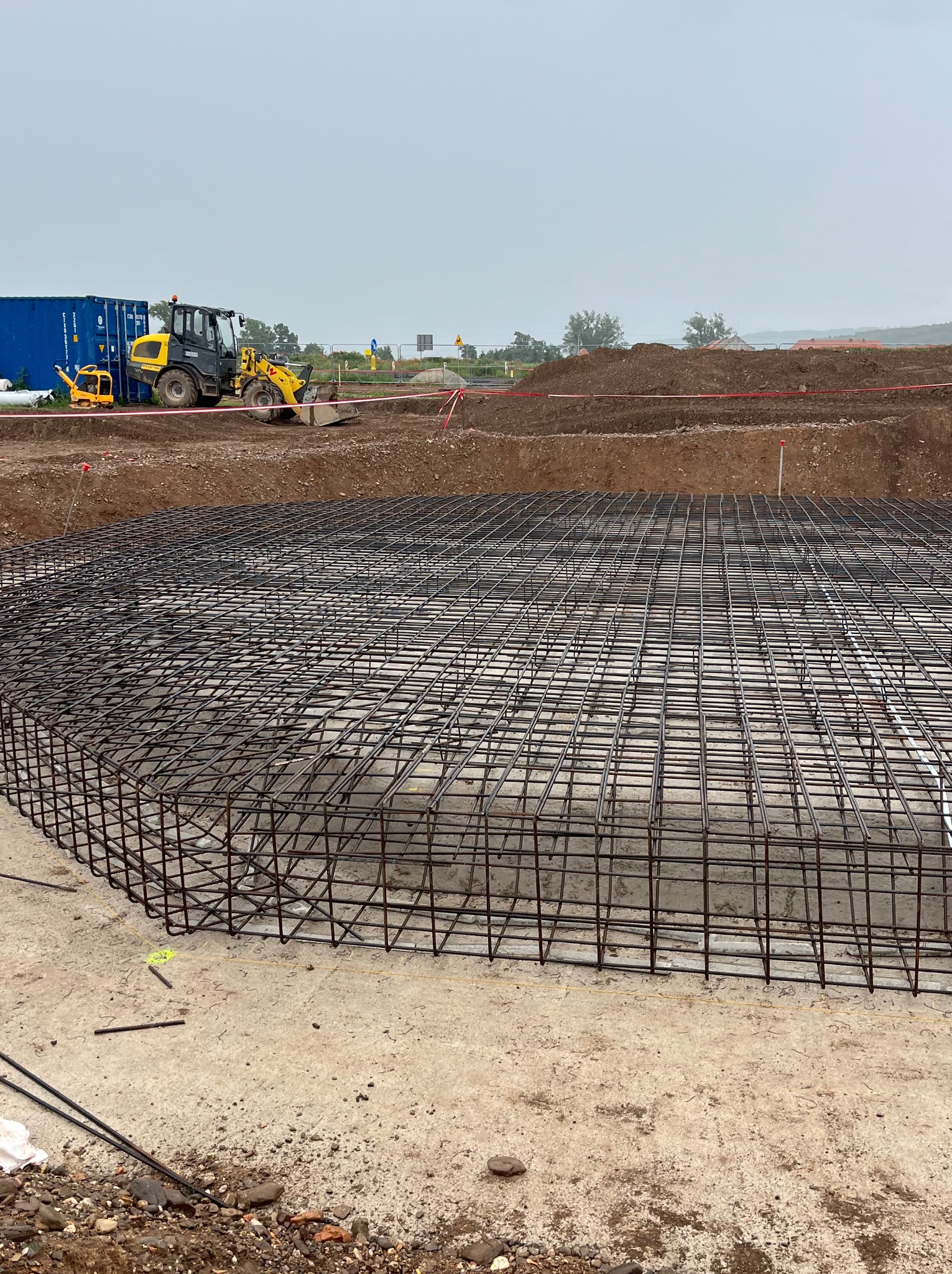 Zbrojenie fundamentu w formie siatki na betonowej płycie, w tle koparka i kontener na budowie. Widok na plac budowy w pochmurny dzień.