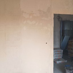 Przemysław Pietrzak - Wnętrze w trakcie prac wykończeniowych: ściany po szpachlowaniu, widoczne worki z gładzią w przejściu, przygotowane do dalszych prac. Minimalistyczny kadr.