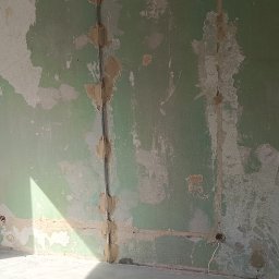 Przemysław Pietrzak - Ściana w trakcie przygotowania do malowania: widoczne ślady po szpachlowaniu, starej farbie i instalacji elektrycznej. W rogu stoi drabina.