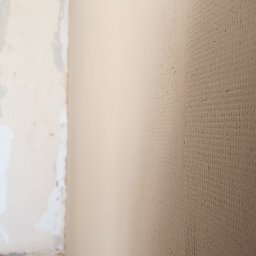 Przemysław Pietrzak - Ściana po szpachlowaniu, gotowa do dalszych prac wykończeniowych. Widoczna faktura gładzi, fragment surowej ściany w tle. Ujęcie z bliska.