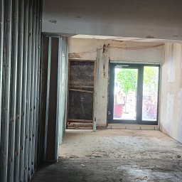 Przemysław Pietrzak - Wnętrze w trakcie prac wykończeniowych: widoczne profile metalowe, ściany przygotowane do malowania, nowe okno balkonowe. Elementy instalacji elektrycznej na ścianie.