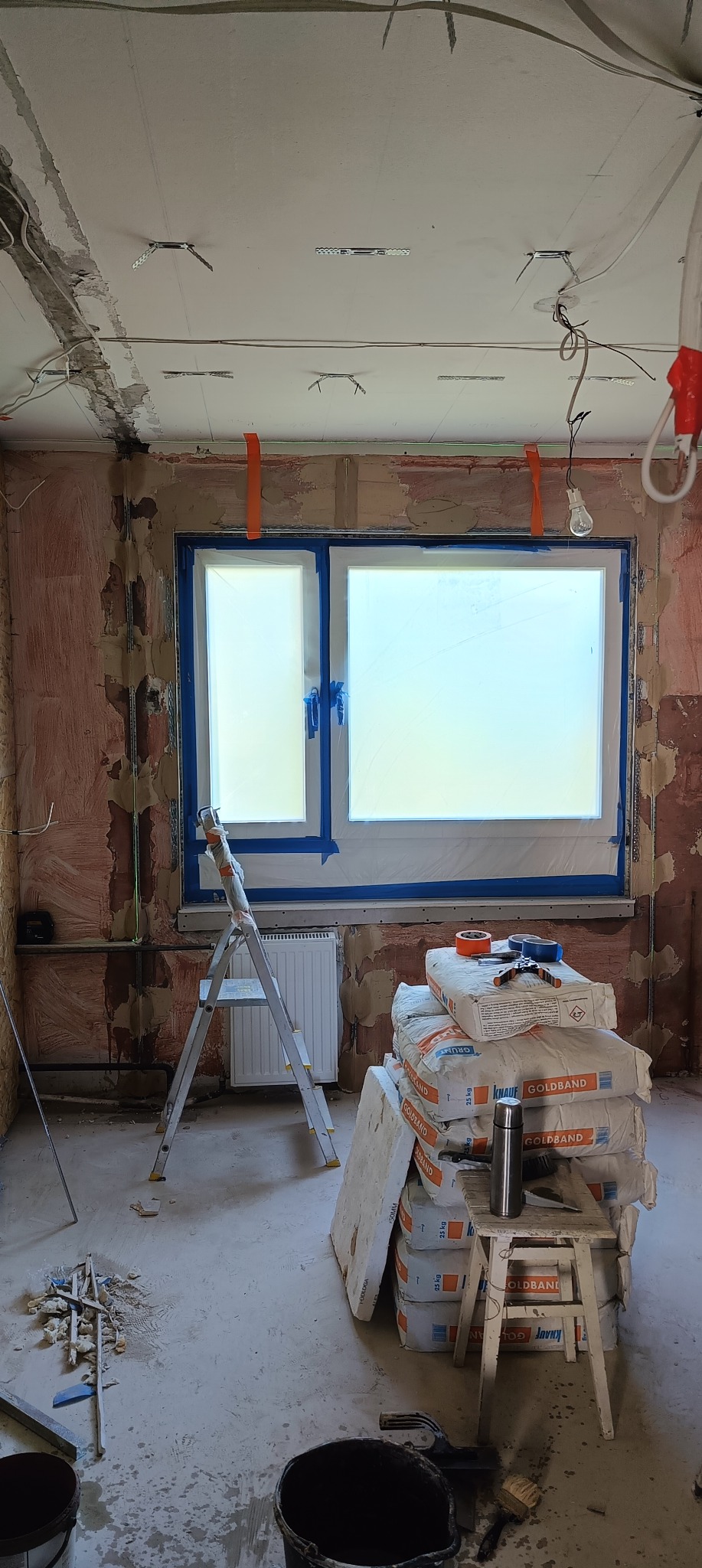 Wnętrze w trakcie remontu: worki Goldband na stołku, drabina przy oknie z niebieską folią, ściany przygotowane do gładzi, widoczne instalacje elektryczne.